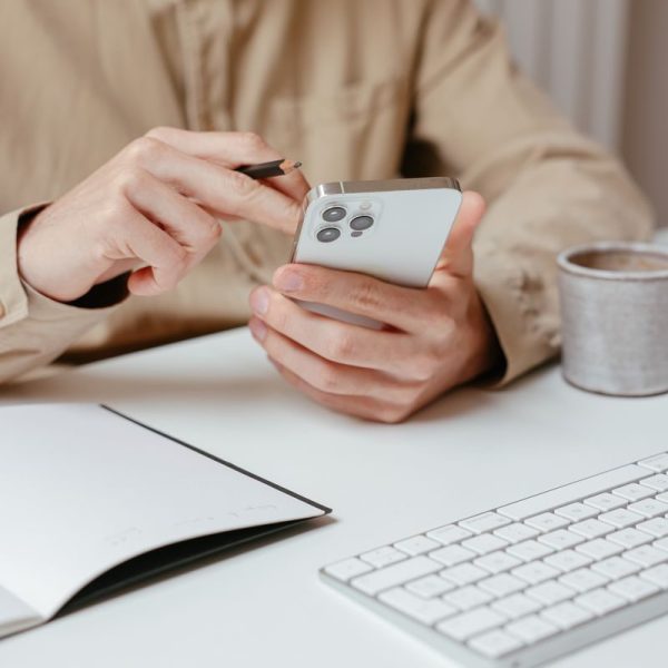Person interacts with smartphone using a stylus at a clean, white office desk with keyboard and notepad.